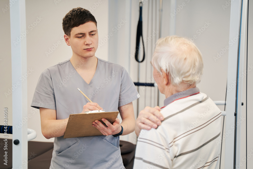Fototapeta premium Attentive physical therapist writing in his notebook about patient complaints