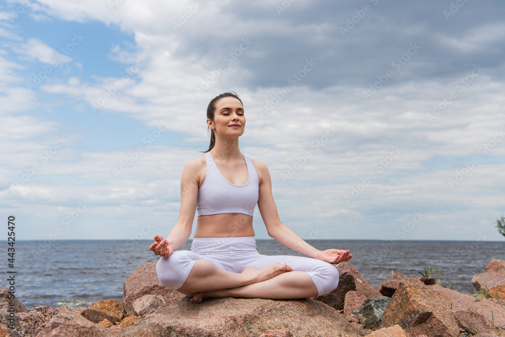 cheerful woman in headphones and sportswear sitting in lotus pose while meditating near sea cheerful woman in headphones and sportswear sitting in lotus pose while meditating near sea