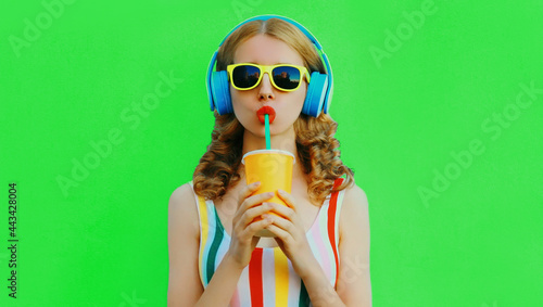 Summer portrait of stylish young woman listening to music in headphones and drink fresh juice on green background