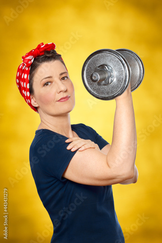 Smiling woman with red kerchief and blue t-shirt lifting a heavy dumbbell in front of yellow background, tribute to american worker icon Rosie the Riveter, strength, power