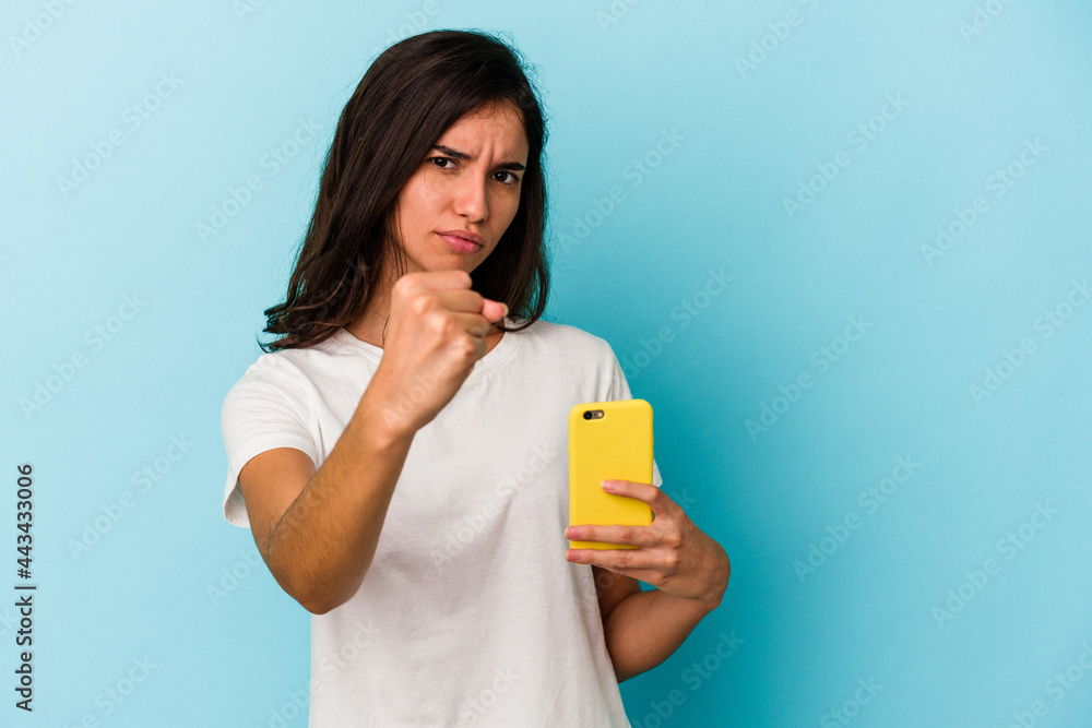 Young caucasian woman holding a mobile phone isolated on blue background showing fist to camera, aggressive facial expression. Young caucasian woman holding a mobile phone isolated on blue background showing fist to camera, aggressive facial expression.
