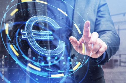 Businessman hand pointing at glowing round euro sign hologram in blurry office interior. Investment and online banking concept. Double exposure.