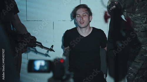 Slowmo tracking shot of armed terrorists taking bag off head of male hostage and forcing him to film confession or ransom message