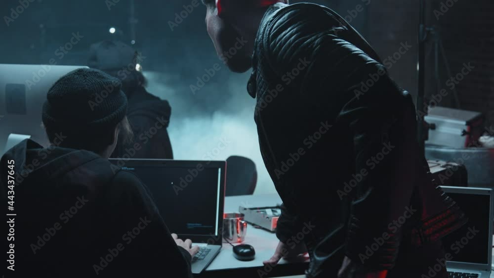Slowmo tracking shot of male hackers in hats talking and coding together on laptop in dark hideout filled with smoke Slowmo tracking shot of male hackers in hats talking and coding together on laptop in dark hideout filled with smoke
