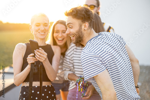 Young stylish friends have fun, showing something on phone and laugh outdoors. Hipsters, millennials hang out at rooftop terrace on sunset photo