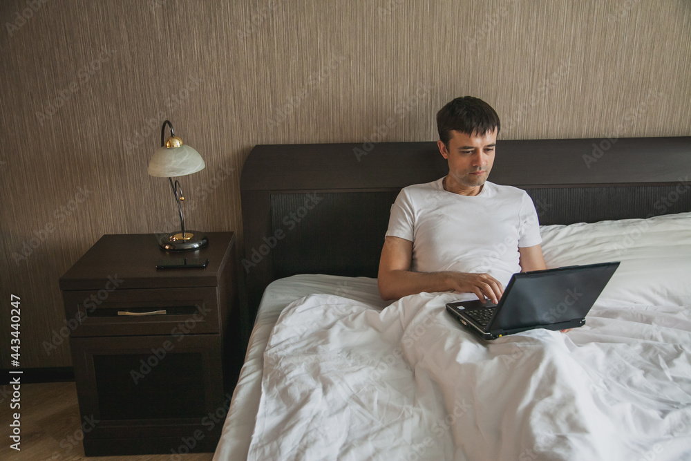 A middle-aged man is sitting in bed in his bedroom and working on a laptop A middle-aged man is sitting in bed in his bedroom and working on a laptop