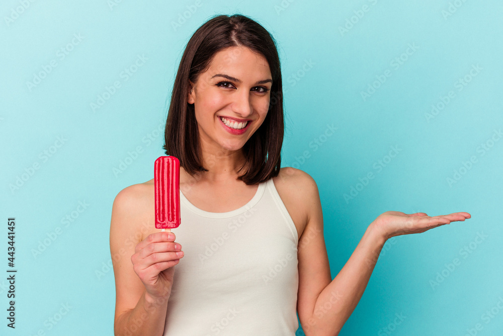 Young caucasian woman holding an ice cream isolated on blue background showing a copy space on a palm and holding another hand on waist. Young caucasian woman holding an ice cream isolated on blue background showing a copy space on a palm and holding another hand on waist.