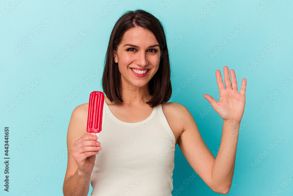 Young caucasian woman holding an ice cream isolated on blue background smiling cheerful showing number five with fingers. Young caucasian woman holding an ice cream isolated on blue background smiling cheerful showing number five with fingers.