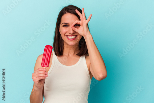 Young caucasian woman holding an ice cream isolated on blue background excited keeping ok gesture on eye.