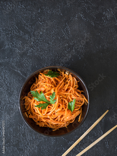 Spicy Asian salad with grated carrots and parsley. On a black background, Korean carrots in a dark bowl with chopsticks. Vegetarian diet. Healthy food concept. View from above