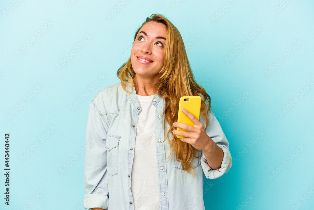 Caucasian woman holding mobile phone isolated on blue background dreaming of achieving goals and purposes Caucasian woman holding mobile phone isolated on blue background dreaming of achieving goals and purposes