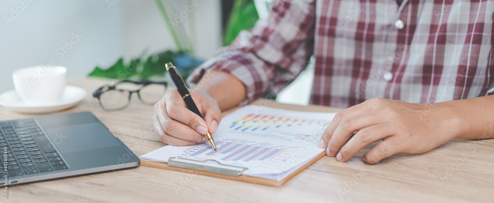 Close-up businessman holding a pen, smartphone and pointing at financial graph checking business report on wooden desk with computer laptop besided at home. Stock photo Close-up businessman holding a pen, smartphone and pointing at financial graph checking business report on wooden desk with computer laptop besided at home. Stock photo