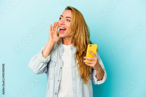 Caucasian woman holding mobile phone isolated on blue background shouting and holding palm near opened mouth.