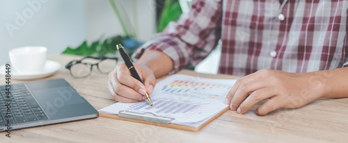 Close-up businessman holding a pen, smartphone and pointing at financial graph checking business report on wooden desk with computer laptop besided at home. Stock photo