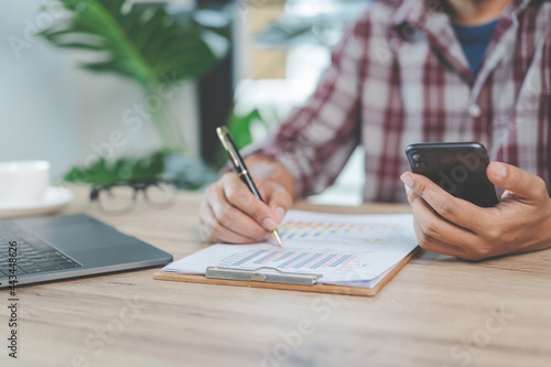 Close-up businessman holding a pen, smartphone and pointing at financial graph checking business report on wooden desk with computer laptop besided at home. Stock photo