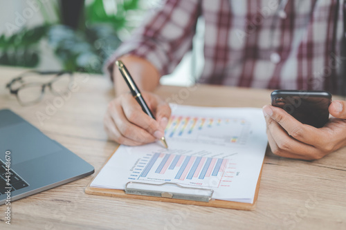 Close-up businessman holding a pen, smartphone and pointing at financial graph checking business report on wooden desk with computer laptop besided at home. Stock photo