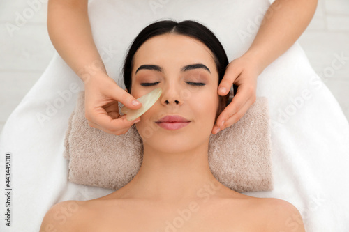 Young woman receiving facial massage with gua sha tool in beauty salon, top view
