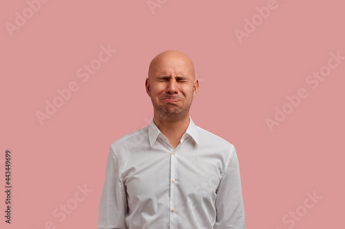 Crying like a child. Desperate bald man has sorrowful expression, cries as a little boy whose parents didnt buy a toy. Have negative emotions, dressed in white shirt. isolated on pink background.