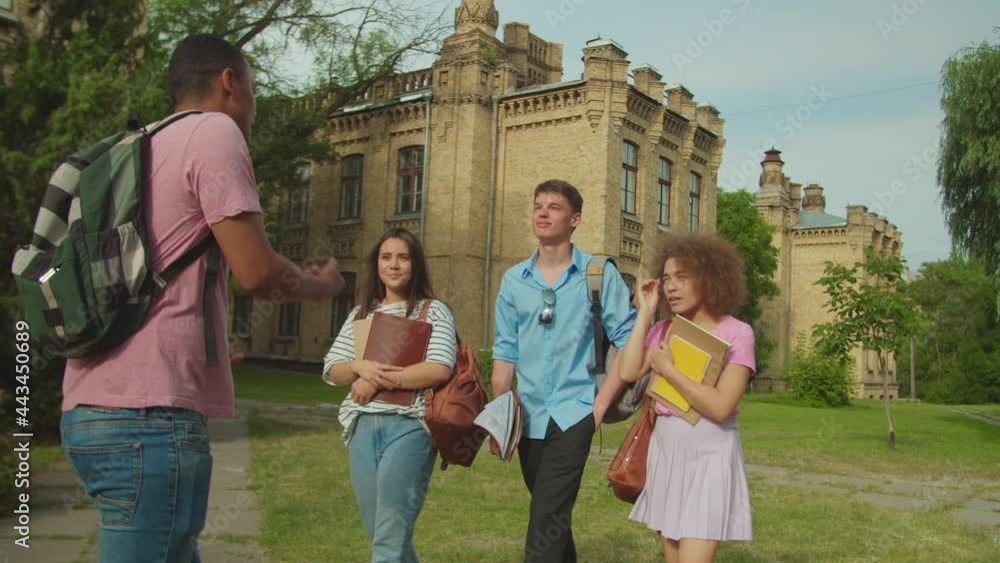Group of positive young friends with bags and copybooks in casual clothing walking along campus building, and listening to one cheerful young friend going backwards with backpack outdoors at sunset