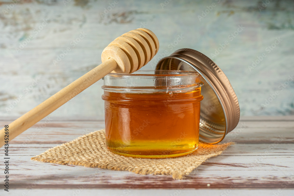 Jar of pure honey from bees with the wooden honey stick resting on the jar on a rustic table. Jar of pure honey from bees with the wooden honey stick resting on the jar on a rustic table.