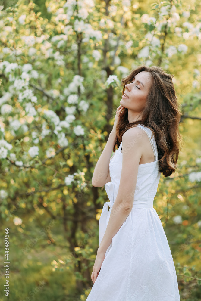 A close-up portrait of a happy pretty gentle smiling young woman with a hairstyle in a white cotton dress having fun walking alone enjoying the smell of blooming flowers in a summer green outdoor park A close-up portrait of a happy pretty gentle smiling young woman with a hairstyle in a white cotton dress having fun walking alone enjoying the smell of blooming flowers in a summer green outdoor park