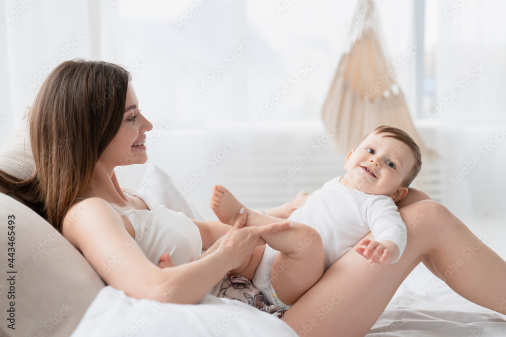 Happy caucasian young single mother playing spending time with little small kid child baby toddler infant newborn at home lying on bed. Motherhood, maternity, postnatal concept Happy caucasian young single mother playing spending time with little small kid child baby toddler infant newborn at home lying on bed. Motherhood, maternity, postnatal concept