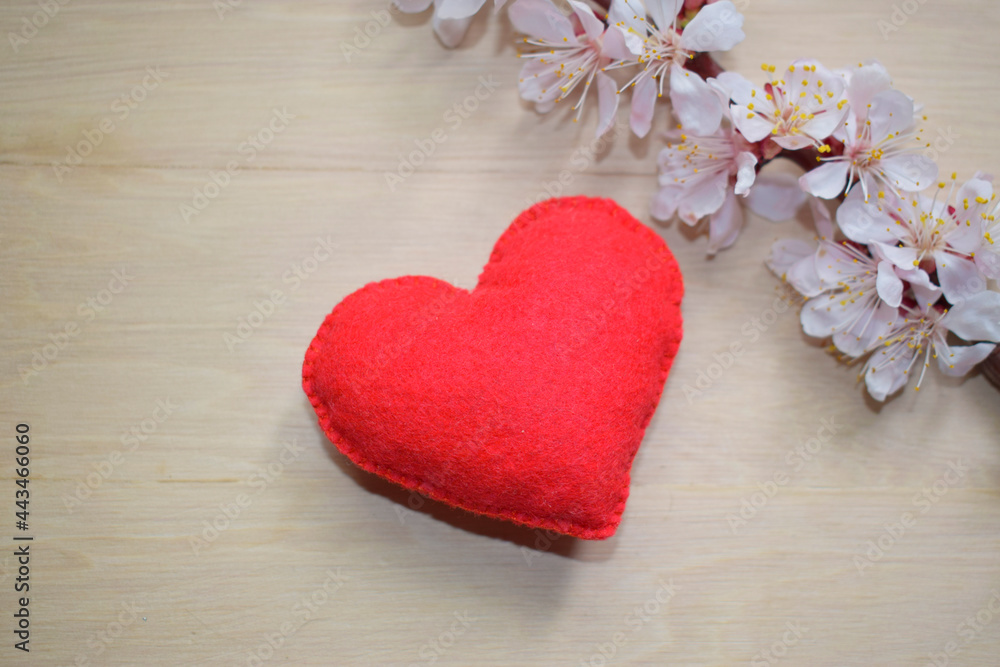 Red felt heart on a wooden surface, next to a blooming twig of a fruit tree. Red felt heart on a wooden surface, next to a blooming twig of a fruit tree.