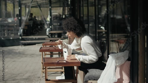 Young business woman eating outdoor having lunch on lunch box sitting at street out of work. Curly haired girl eat noodles with chopsticks while having lunch time outdoors photo