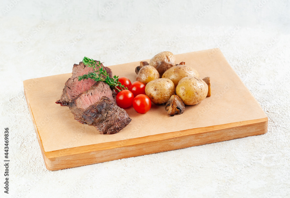 Veal steak with baked potatoes and mushrooms. Decorated with herbs and cherry tomatoes. Served on a wooden board. Close-up. Veal steak with baked potatoes and mushrooms. Decorated with herbs and cherry tomatoes. Served on a wooden board. Close-up.