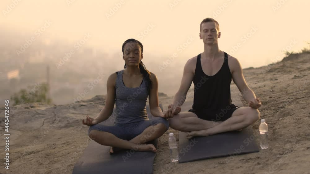Young multiethnic friends in sport outfit sitting in lotus position with eyes closed and meditating on yoga mats outdoors. Concept of yoga, breathing practice and peacefulness. Young multiethnic friends in sport outfit sitting in lotus position with eyes closed and meditating on yoga mats outdoors. Concept of yoga, breathing practice and peacefulness.