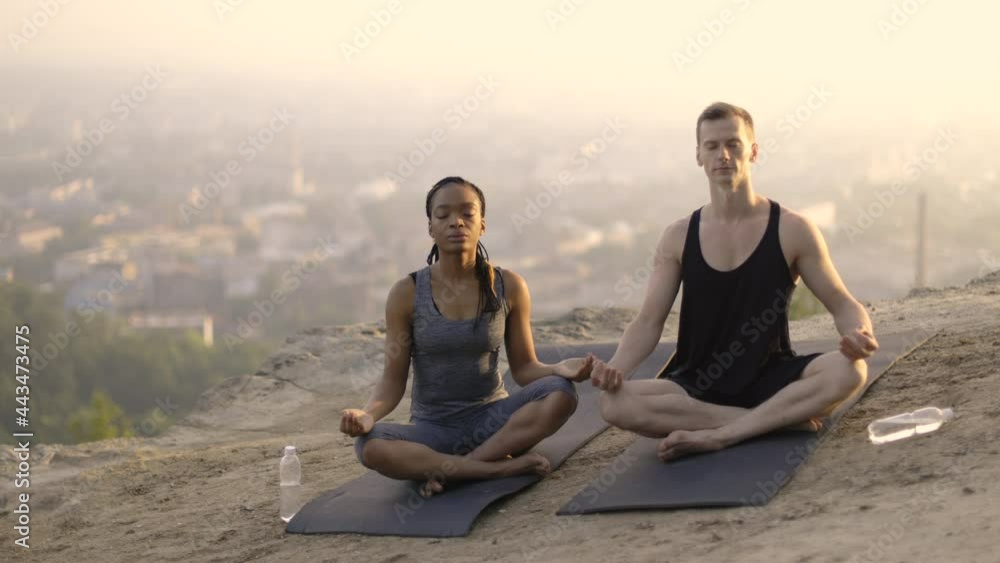 Two multiethnic friends, woman and man, in sport outfit sitting in lotus position with eyes closed and meditating on yoga mats outdoors. Concept of yoga, breathing practice and peacefulness. Two multiethnic friends, woman and man, in sport outfit sitting in lotus position with eyes closed and meditating on yoga mats outdoors. Concept of yoga, breathing practice and peacefulness.