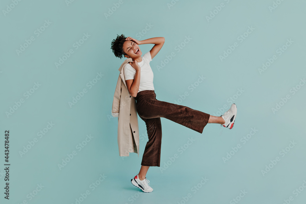 Naklejka premium Excited african american woman in pants dancing on turquoise background. Full length view of adorable black girl holding trench coat.
