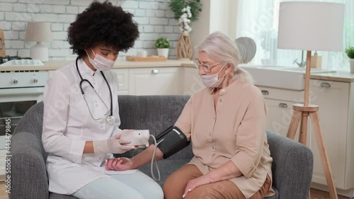 Afro american woman doctor in mask checks blood pressure senier woman at home sitting on couch. Family Doctor, Patient Support, Help at Home, Caring for the Sick. photo