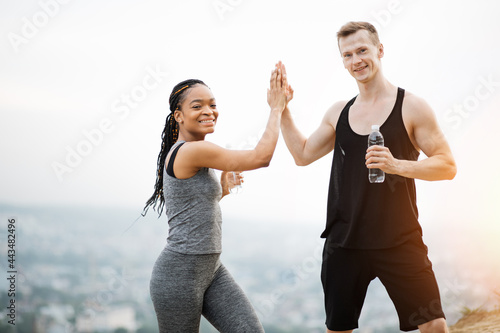 Multicultural couple wearing sport clothes posing with bottle of water in hands outdoors. Caucasian man and black woman giving high five. Active and healthy lifestyles.