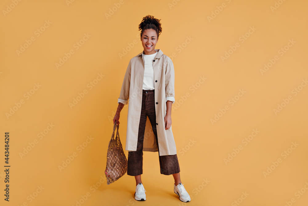 Fototapeta premium Full length view of confident african american woman with string bag. Studio shot of black girl wearing beige trench coat.