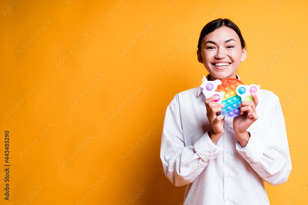Happy asian girl in shirt holding childrens toys pop it and simple dimple smiling broadly while standing on yellow background Happy asian girl in shirt holding childrens toys pop it and simple dimple smiling broadly while standing on yellow background