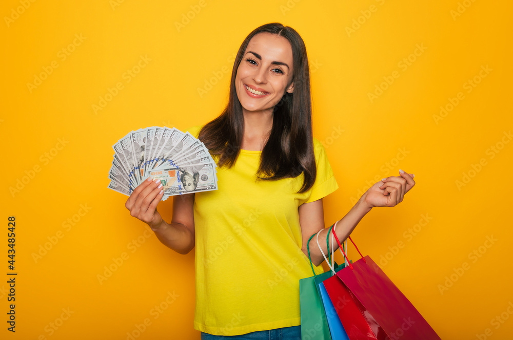 Modern cute smiling young woman with shopping bags and dollars in her hands is posing over yellow wall Modern cute smiling young woman with shopping bags and dollars in her hands is posing over yellow wall