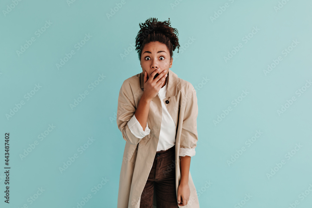 Shocked african american woman in trench coat covering mouth with hand. Front view of black girl expressing amazement on turquoise background. Shocked african american woman in trench coat covering mouth with hand. Front view of black girl expressing amazement on turquoise background.