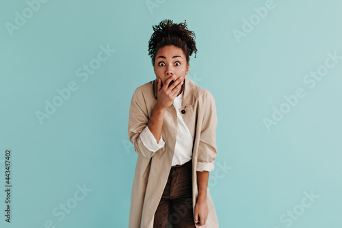 Shocked african american woman in trench coat covering mouth with hand. Front view of black girl expressing amazement on turquoise background.