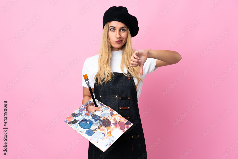 Young artist woman holding a palette over isolated pink background showing thumb down sign