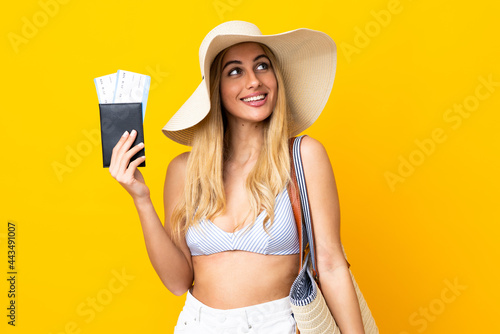 Young Uruguayan blonde woman in swimsuit holding a passport over isolated yellow background looking up while smiling