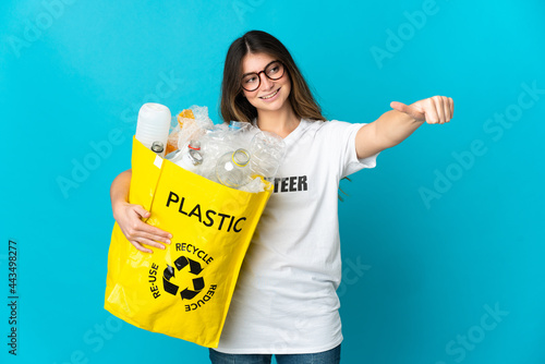 Woman holding a bag full of bottles to recycle isolated on blue background giving a thumbs up gesture