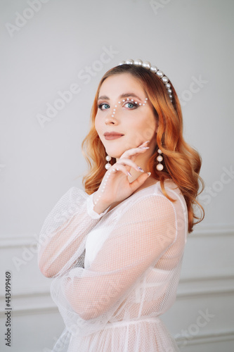 portrait of a beautiful young woman with red hair in a white dress and makeup made of pearls on her face, hand. 