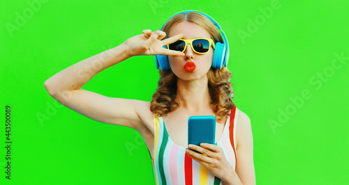 Summer colorful portrait of stylish young woman listening to music in headphones with phone on green background