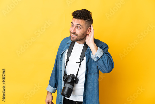 Photographer man isolated on yellow background listening to something by putting hand on the ear
