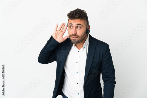 Telemarketer man working with a headset isolated on white background listening to something by putting hand on the ear