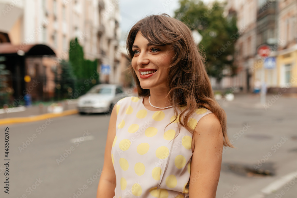 Smiling attractive girl with wavy brown hair wearing summer bright dress walking across the street in summer warm day