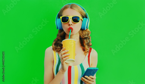 Summer colorful portrait of stylish young woman drinking fresh juice listening to music in headphones with smartphone on green background