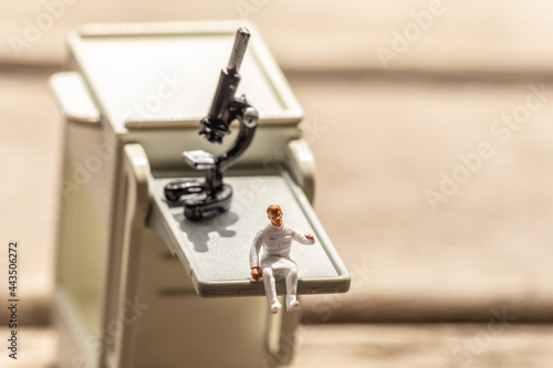 A human figurine sitting on a medical trolley , medical staff figurine with a microscope in the background to symbolize lab process