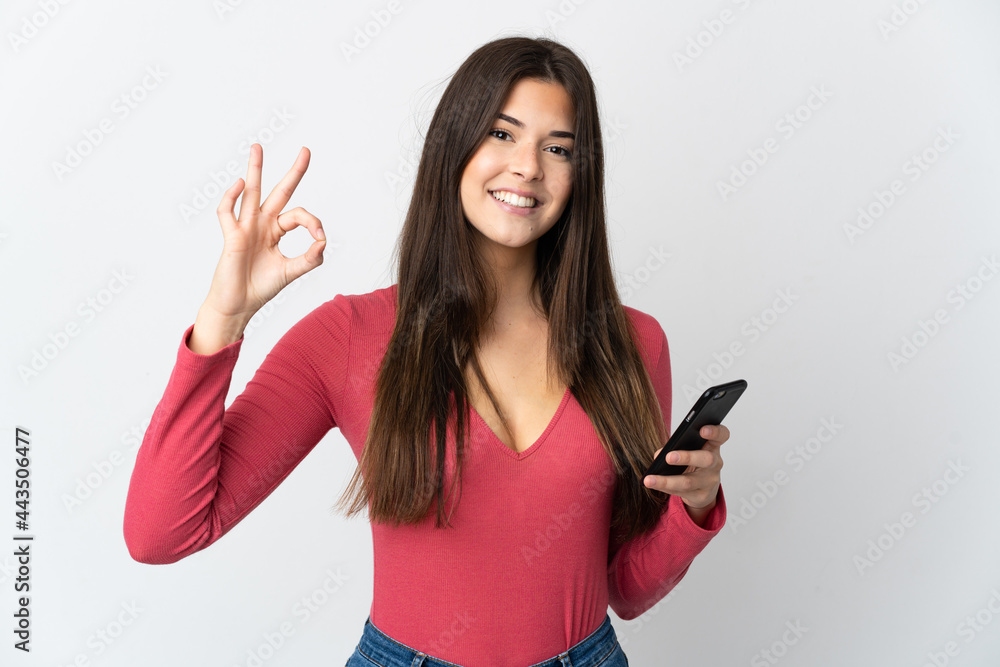 Teenager Brazilian girl isolated on white background using mobile phone and doing OK sign Teenager Brazilian girl isolated on white background using mobile phone and doing OK sign
